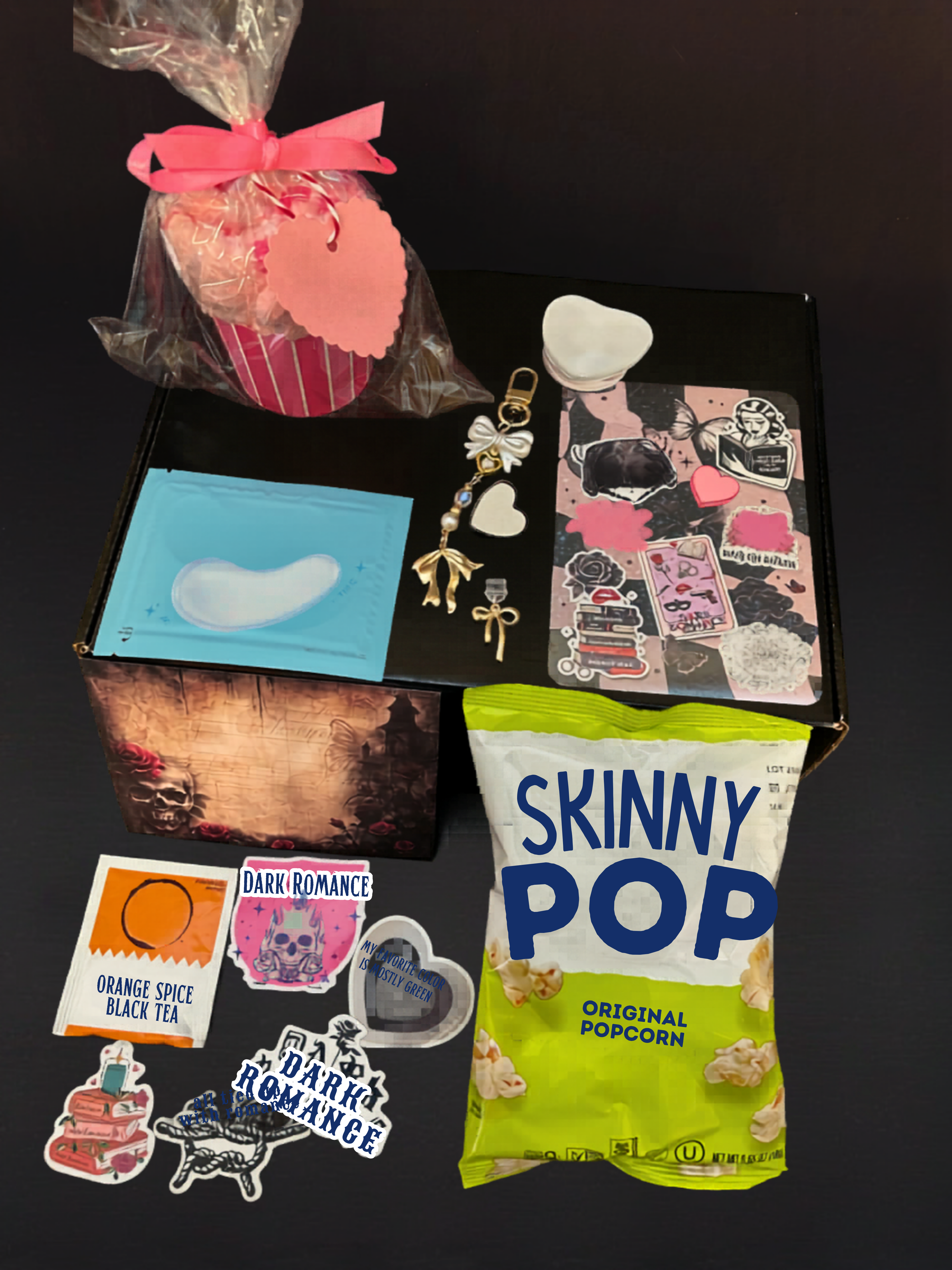 Assorted items including a box, stickers, a bag of Skinny Pop popcorn, and a small pink cupcake on a dark surface.